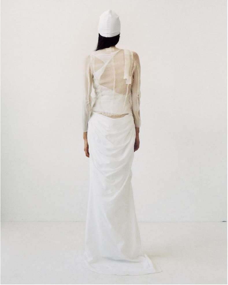 A woman is seen from the back wearing a white cap, a sheer long-sleeved shirt, and a draped white skirt against a white background.