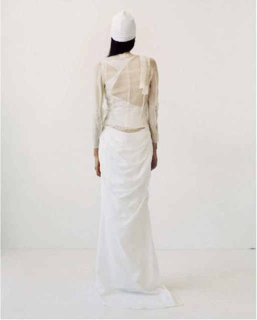 A woman is seen from the back wearing a white cap, a sheer long-sleeved shirt, and a draped white skirt against a white background.