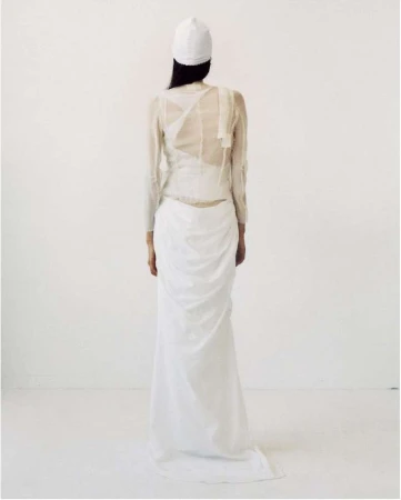 A woman is seen from the back wearing a white cap, a sheer long-sleeved shirt, and a draped white skirt against a white background.