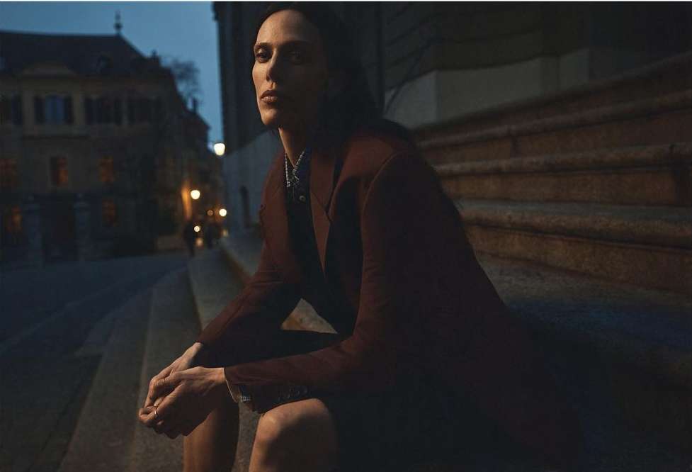 Adult woman in dark coat sitting on steps outside at twilight in an urban setting, city buildings in the background and soft, diffused lighting.
