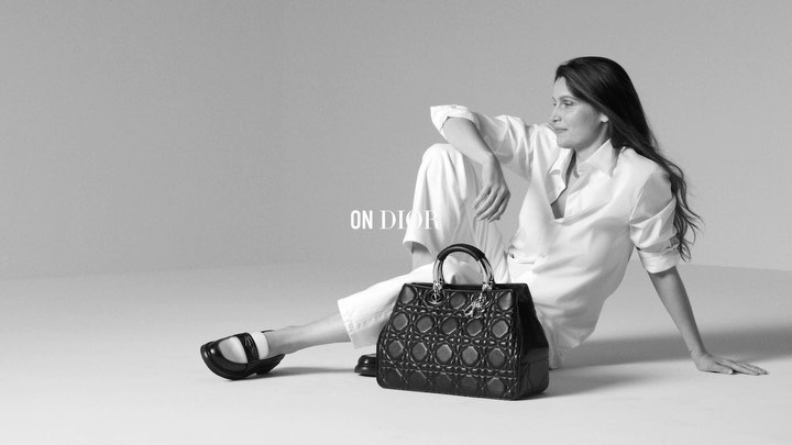 Elegant studio shot of a woman in a white outfit seated next to a black leather Dior handbag with metal hardware on a grey backdrop.