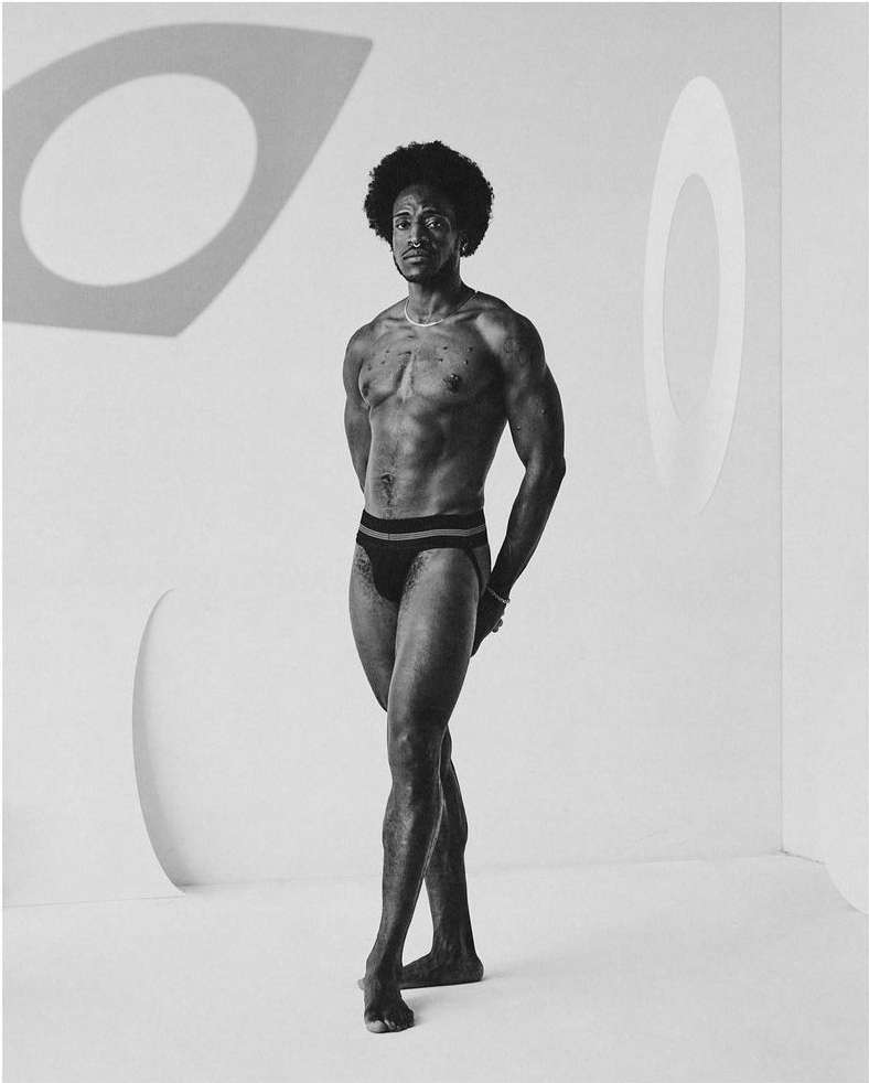 Monochrome studio shot of a muscular adult male posing against a white wall with geometric shadows, wearing a swimsuit.