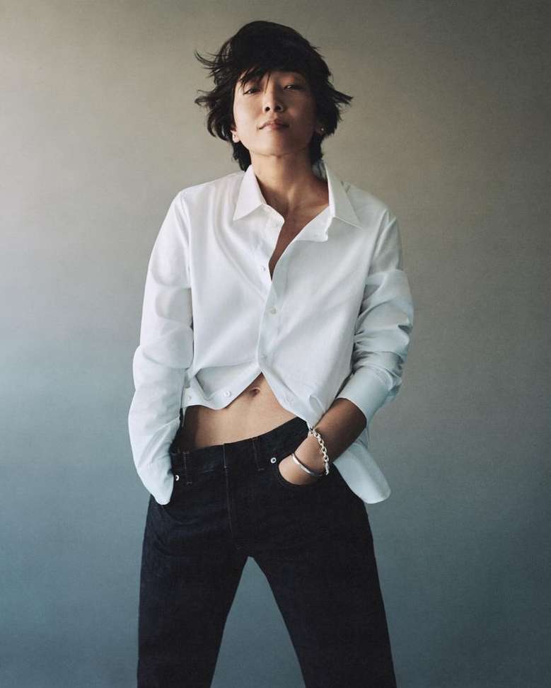 An adult posing in a white button-down shirt and dark jeans with a light gray backdrop. Indoor fashion shot with natural lighting.