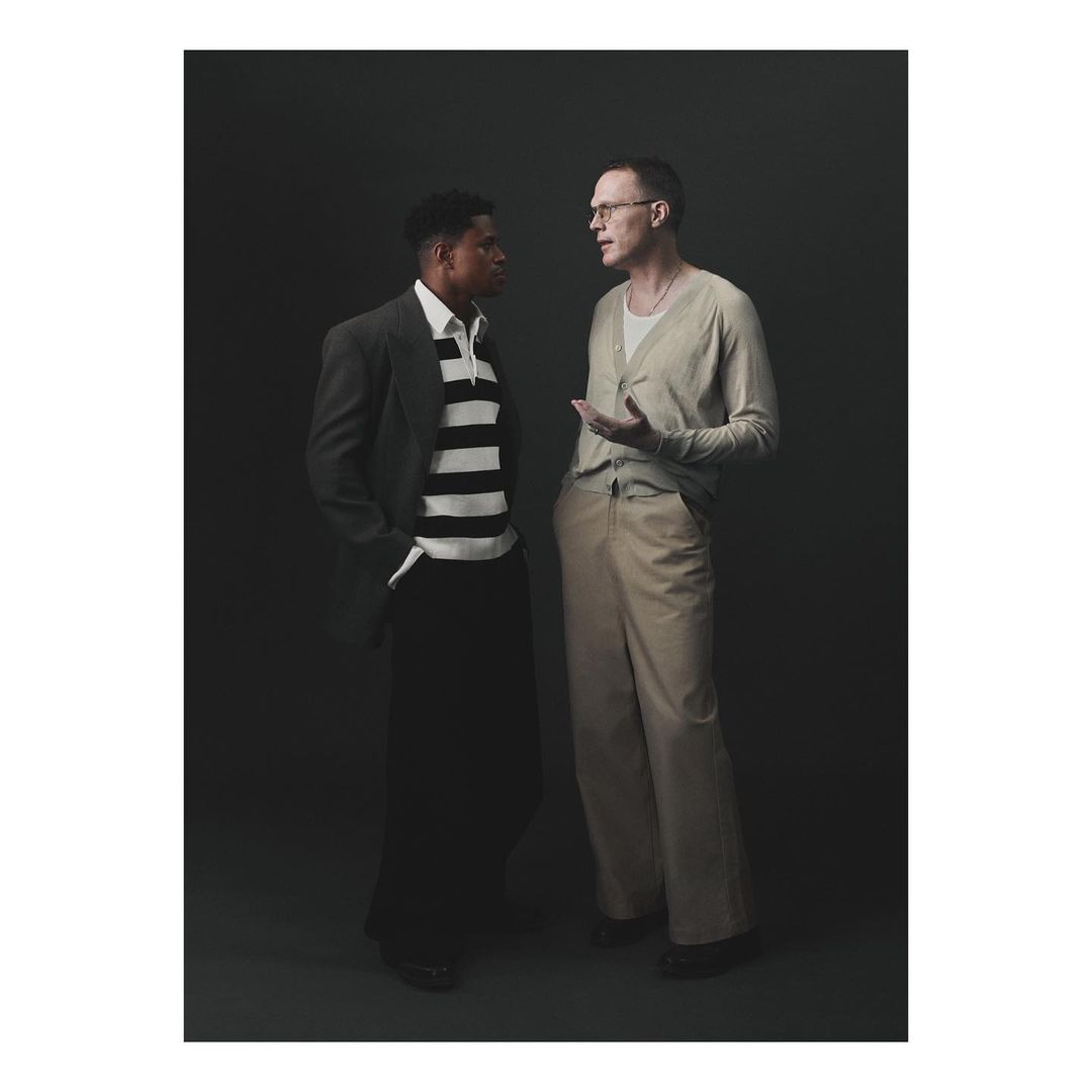 Two men stand in a photo studio. One wears a striped shirt and jacket, while the other wears a cardigan and glasses.