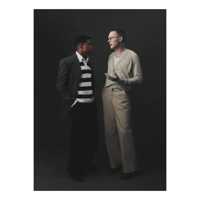 Two men stand in a photo studio. One wears a striped shirt and jacket, while the other wears a cardigan and glasses.