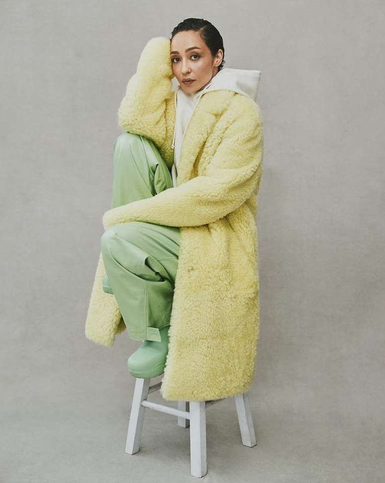 A woman in a yellow coat sits on a white stool, posing in a studio with a neutral background, wearing a hoodie, pants, and boots.