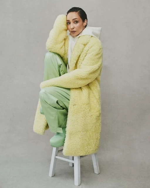 A woman in a yellow coat sits on a white stool, posing in a studio with a neutral background, wearing a hoodie, pants, and boots.