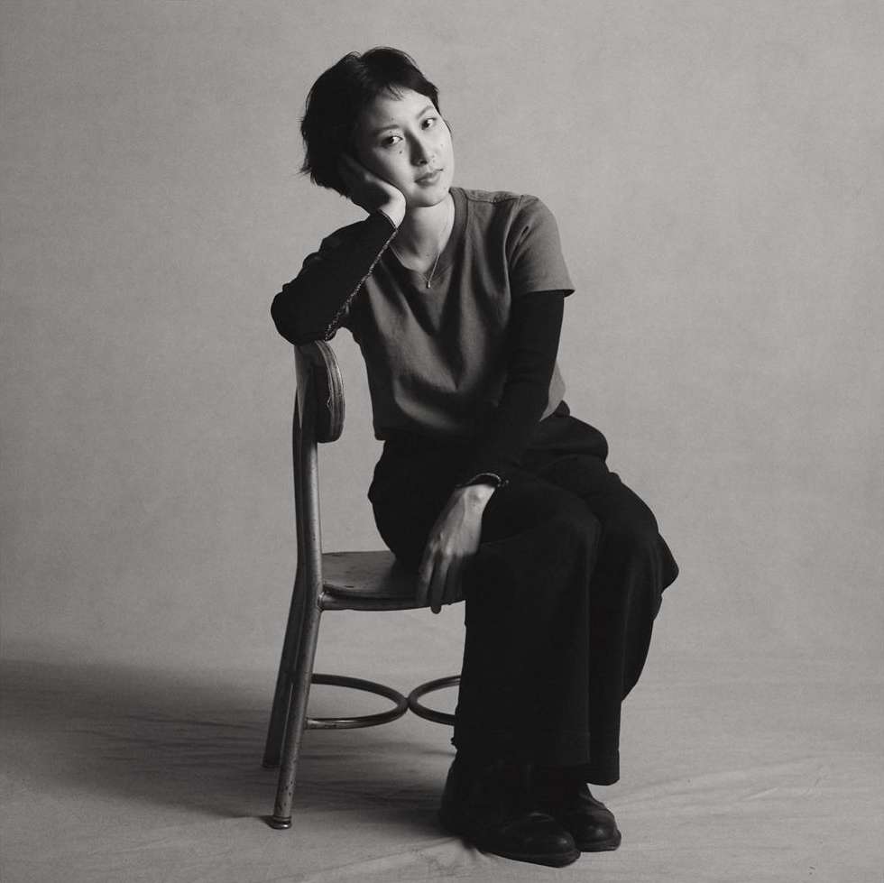 Portrait of a young Asian woman with short hair sitting on a wooden chair in a photo studio, wearing a t-shirt and wide-legged pants.