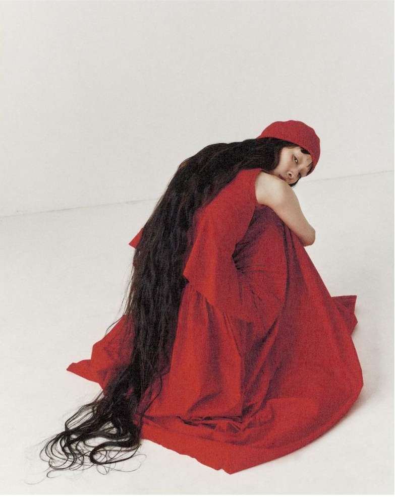 Elegant adult woman with long dark hair sits in a draped red gown and matching headscarf against a minimalist light background in a studio.