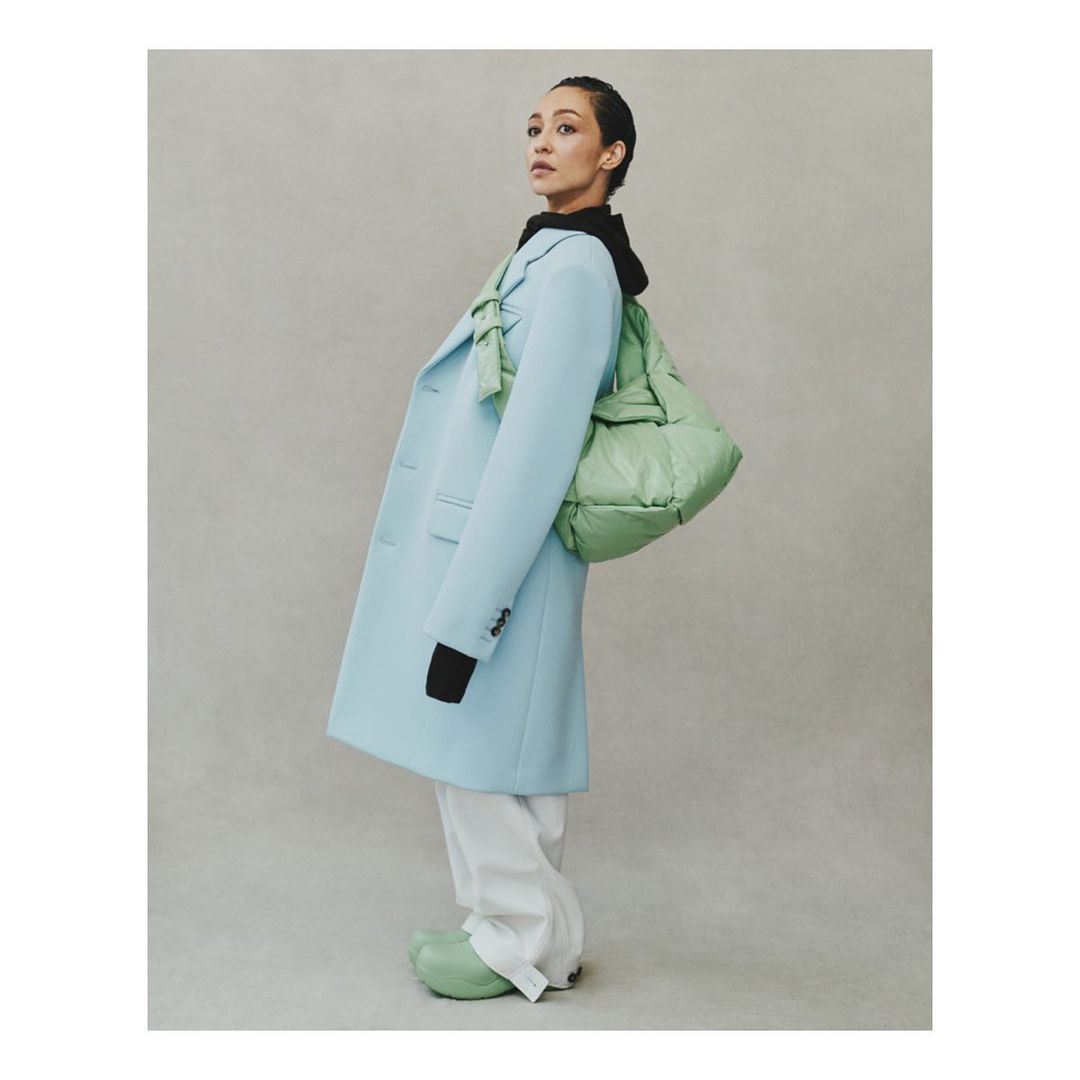 A woman in a light blue coat and white pants poses with a light green padded bag in front of a neutral-toned backdrop.