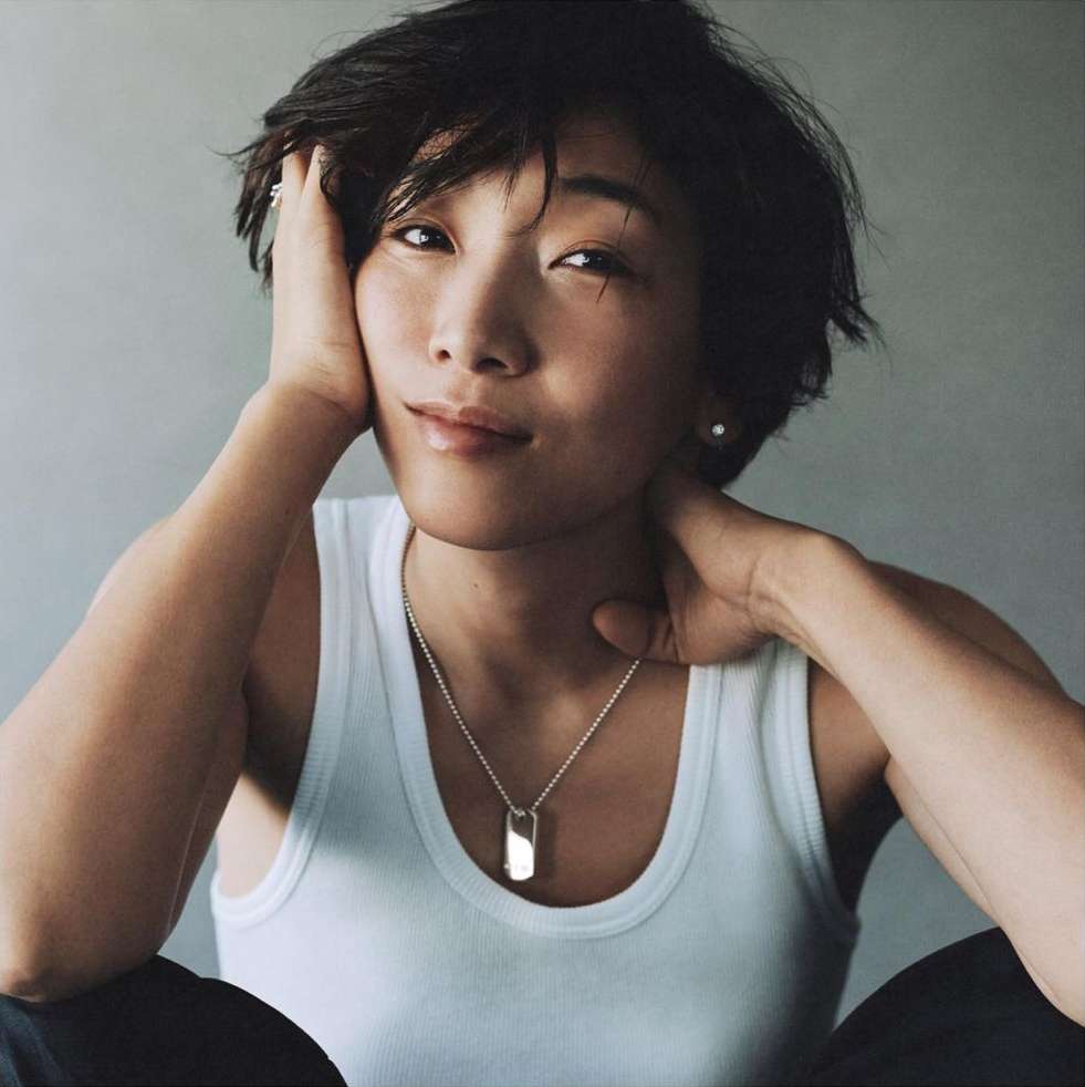 Asian woman in a white tank top with short hair poses with her hands near her face, looking at the camera in a portrait shot.