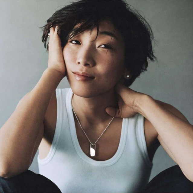 Asian woman in a white tank top with short hair poses with her hands near her face, looking at the camera in a portrait shot.