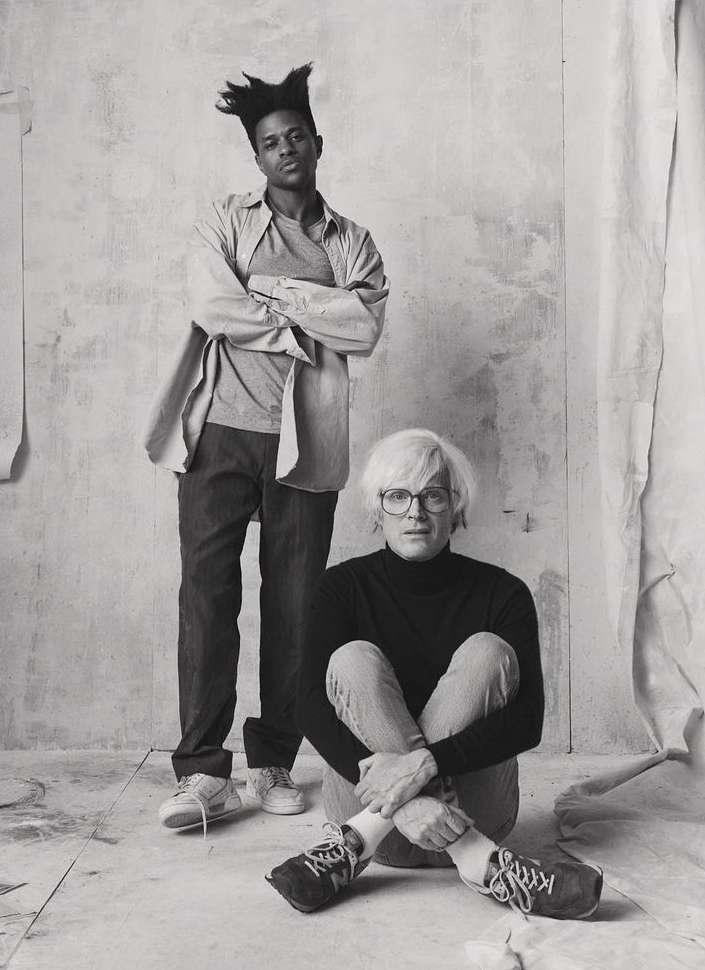 Black and white portrait of two adult males, one standing with arms crossed, the other seated on the floor with knees up.