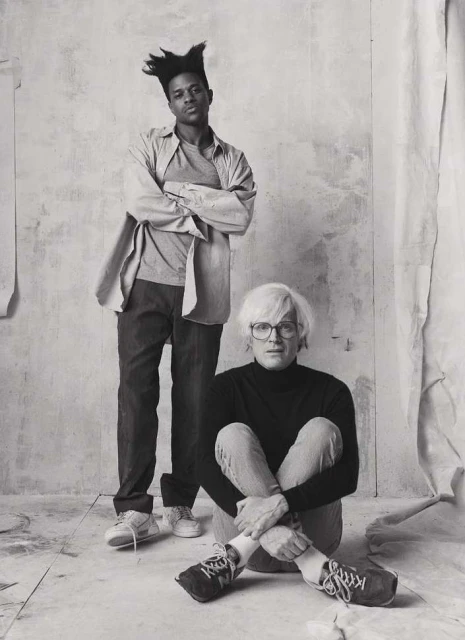 Black and white portrait of two adult males, one standing with arms crossed, the other seated on the floor with knees up.