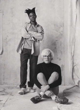 Black and white portrait of two adult males, one standing with arms crossed, the other seated on the floor with knees up.