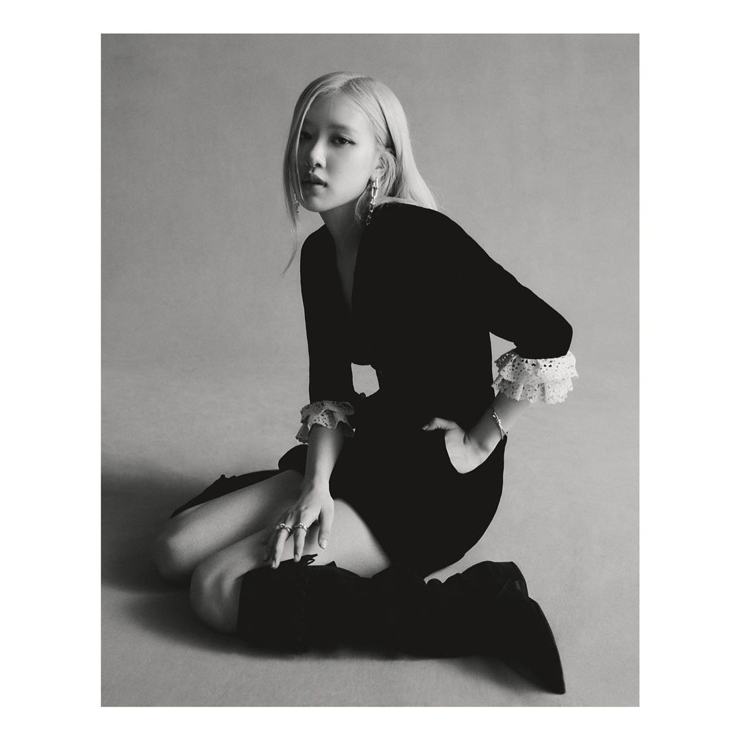 Black and white portrait of a blonde woman kneeling in a black dress with lace cuffs and high black boots, posing and looking at the camera.