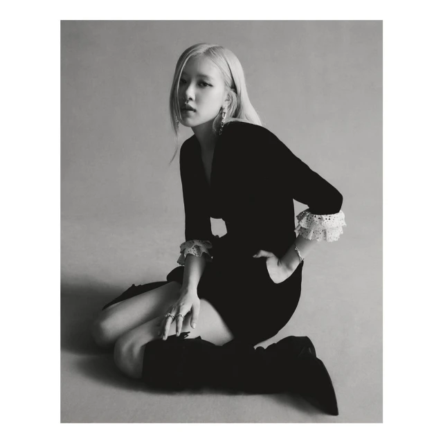 Black and white portrait of a blonde woman kneeling in a black dress with lace cuffs and high black boots, posing and looking at the camera.