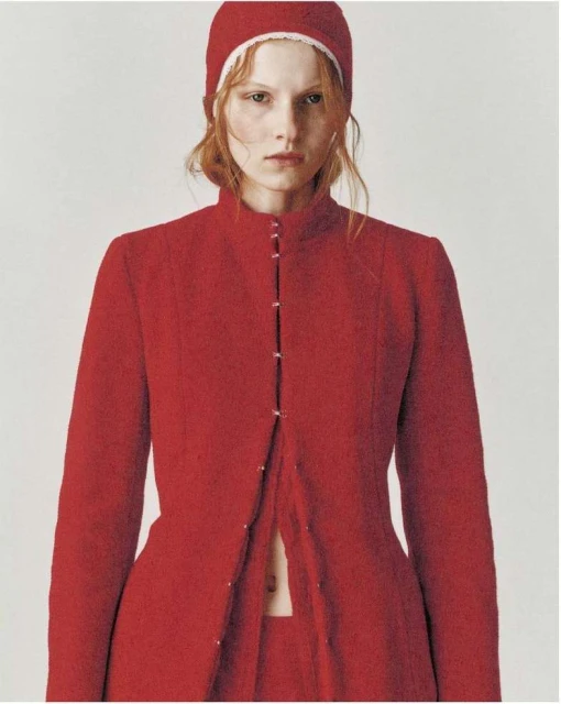 A red-haired model in a red knit cap and tailored red coat poses in a studio with soft lighting for a fashion editorial.