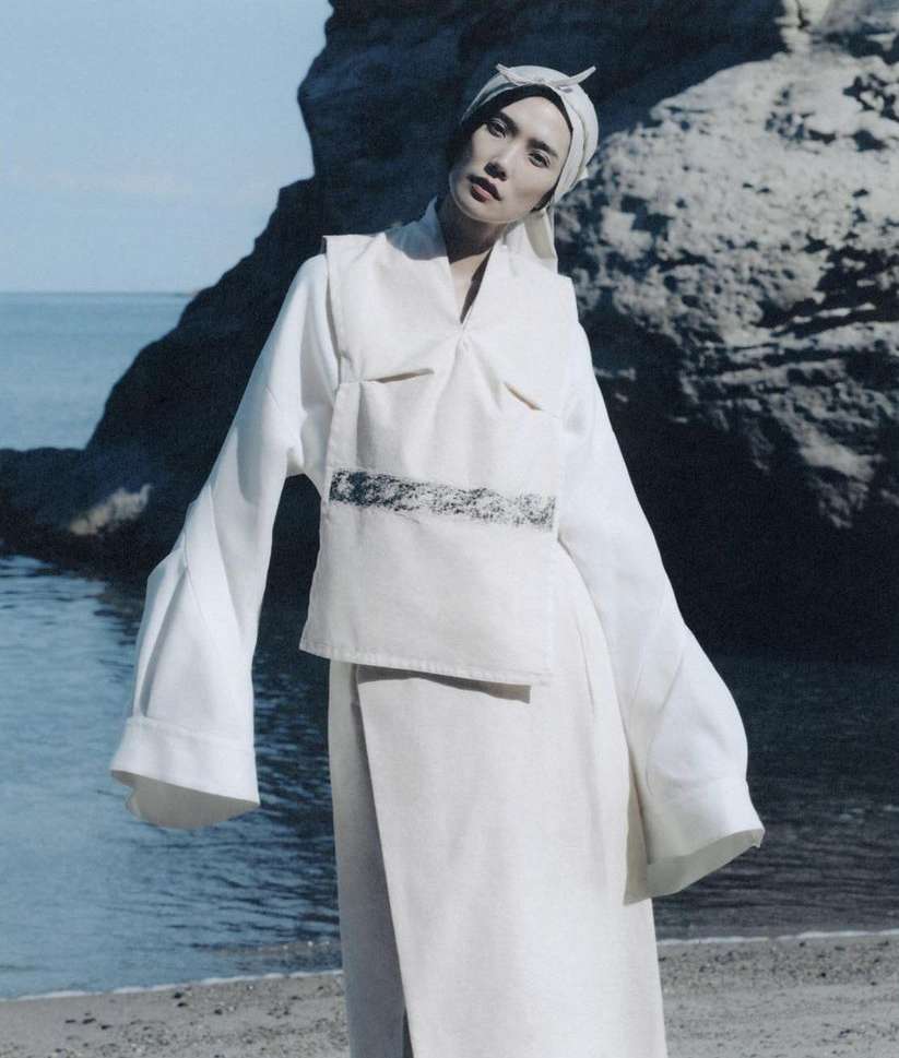 A woman in a white outfit poses on a sandy beach, set against a backdrop of rock formations and the sea, showcasing a blend of fashion and nature.