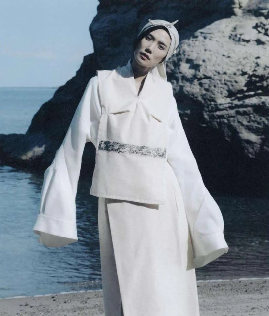 A woman in a white outfit poses on a sandy beach, set against a backdrop of rock formations and the sea, showcasing a blend of fashion and nature.