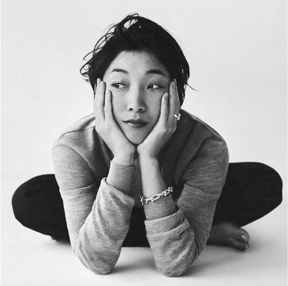 Black-and-white portrait of an Asian woman with short hair, resting her face in her hands while kneeling in a photo studio.