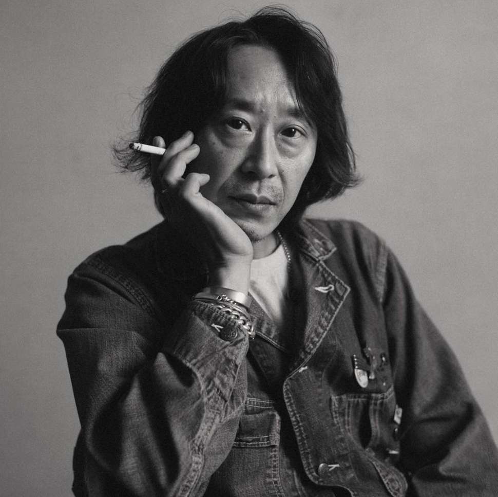 Black and white portrait of an Asian man holding a cigarette. He's wearing a denim jacket and silver bracelets against a neutral background.