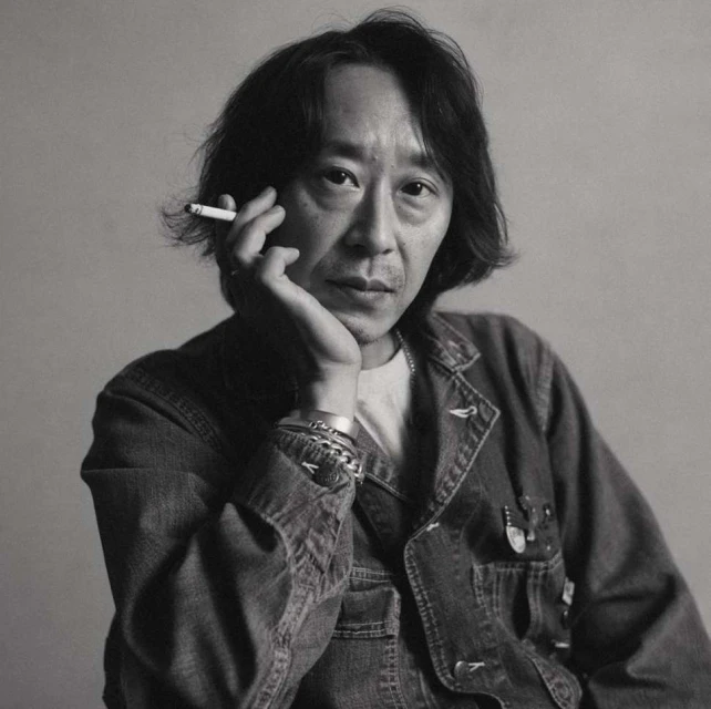 Black and white portrait of an Asian man holding a cigarette. He's wearing a denim jacket and silver bracelets against a neutral background.