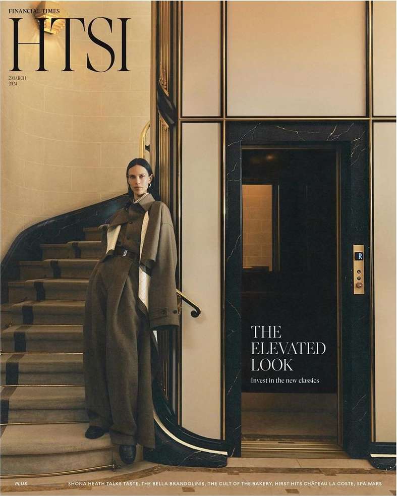 Fashion editorial shot of a stylish adult female in an olive trench coat and wide-leg pants, posing indoors on a marble staircase.