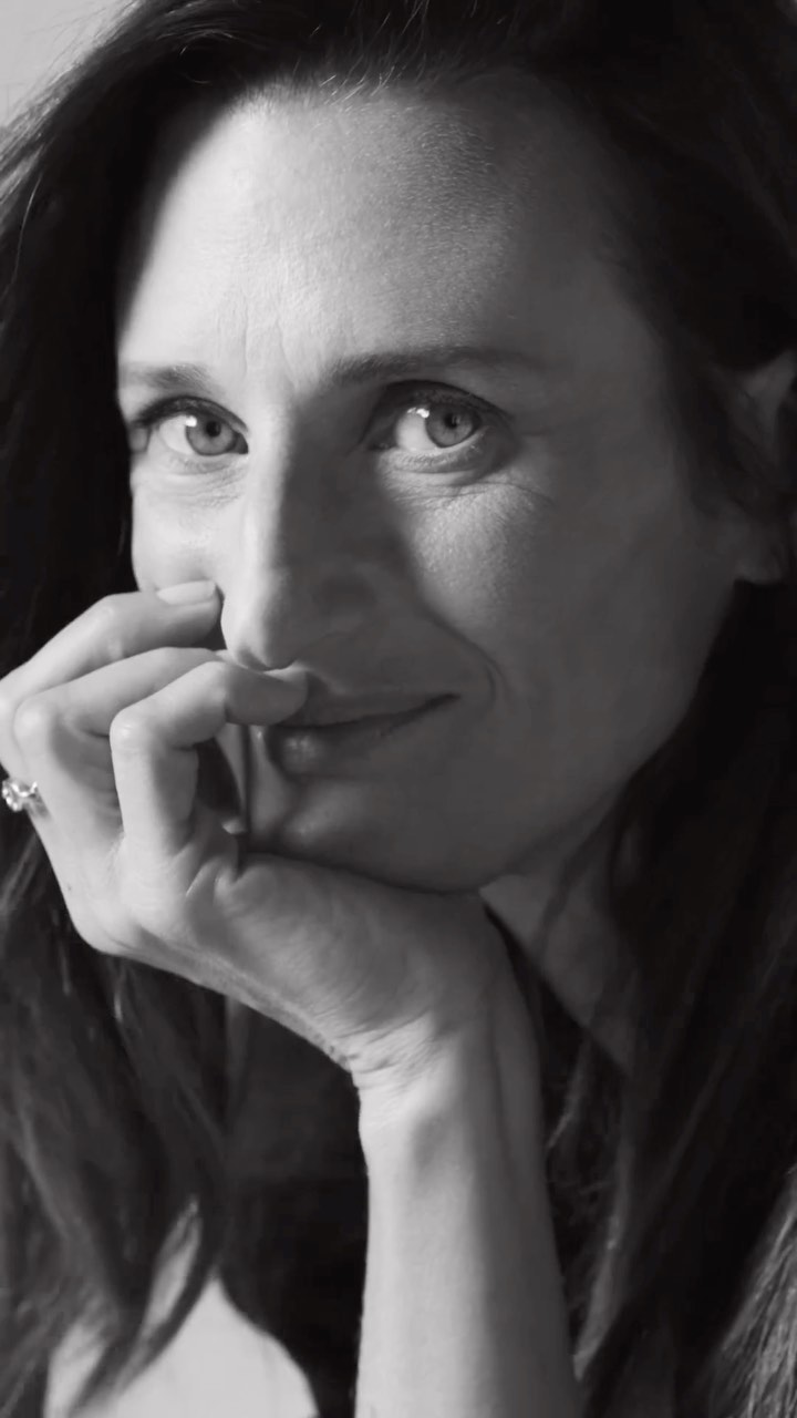 Monochrome portrait of a woman with dark hair resting her face on her hand, smiling softly at eye level in an indoor setting.