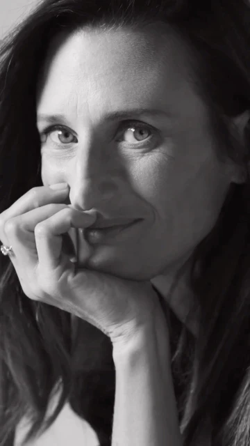 Monochrome portrait of a woman with dark hair resting her face on her hand, smiling softly at eye level in an indoor setting.