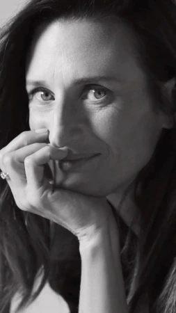 Monochrome portrait of a woman with dark hair resting her face on her hand, smiling softly at eye level in an indoor setting.