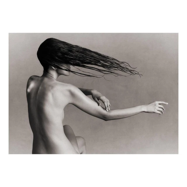 Black and white studio shot of an adult female with long dark hair cascading down her back, arm extended, against a neutral background.