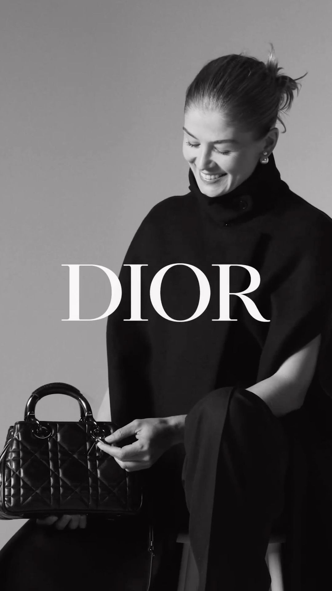 Black and white portrait of a smiling adult woman in a dark outfit, seated and holding a black Lady Dior handbag.
