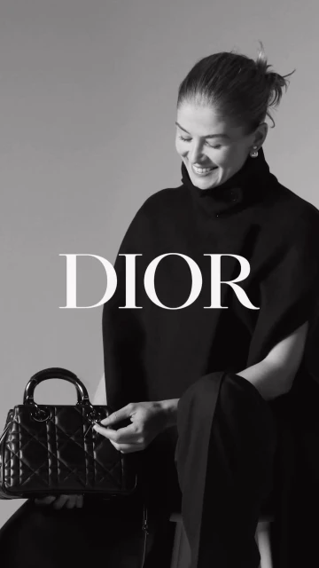 Black and white portrait of a smiling adult woman in a dark outfit, seated and holding a black Lady Dior handbag.