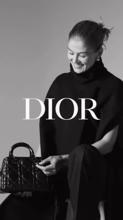 Black and white portrait of a smiling adult woman in a dark outfit, seated and holding a black Lady Dior handbag.