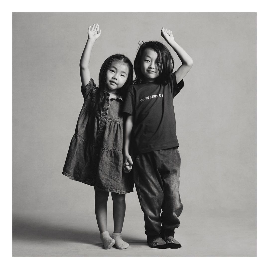 Two young Asian children, a girl in a denim dress and a boy in a T-shirt and jeans, stand side-by-side holding hands and raising arms.