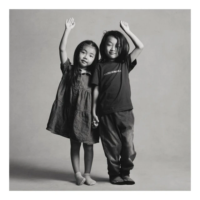 Two young Asian children, a girl in a denim dress and a boy in a T-shirt and jeans, stand side-by-side holding hands and raising arms.