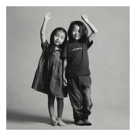 Two young Asian children, a girl in a denim dress and a boy in a T-shirt and jeans, stand side-by-side holding hands and raising arms.