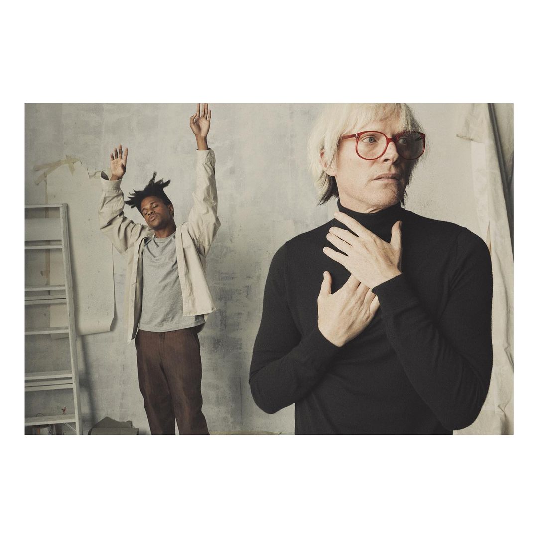 Two people in an indoor photo studio setting: a black male with hands raised and a white male in red glasses and black turtleneck.