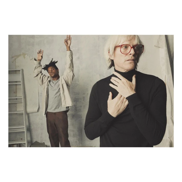 Two people in an indoor photo studio setting: a black male with hands raised and a white male in red glasses and black turtleneck.