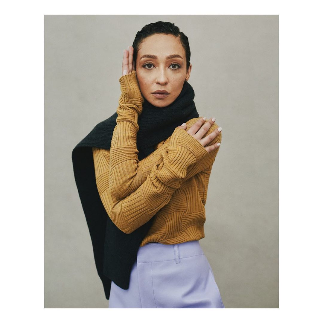 Portrait of adult woman in a golden sweater and black scarf posing with hands near her face in a photo studio setting.