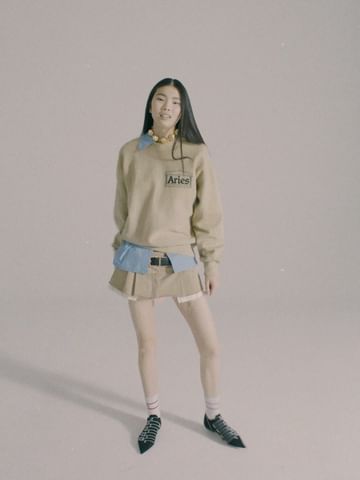 A woman in a beige sweatshirt, short skirt, and patterned shoes poses in a neutral studio setting, fashion forward and confidently stylish.