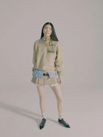 A woman in a beige sweatshirt, short skirt, and patterned shoes poses in a neutral studio setting, fashion forward and confidently stylish.