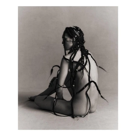 Seated woman in profile with long braided hair draped across her back against a neutral grey studio background