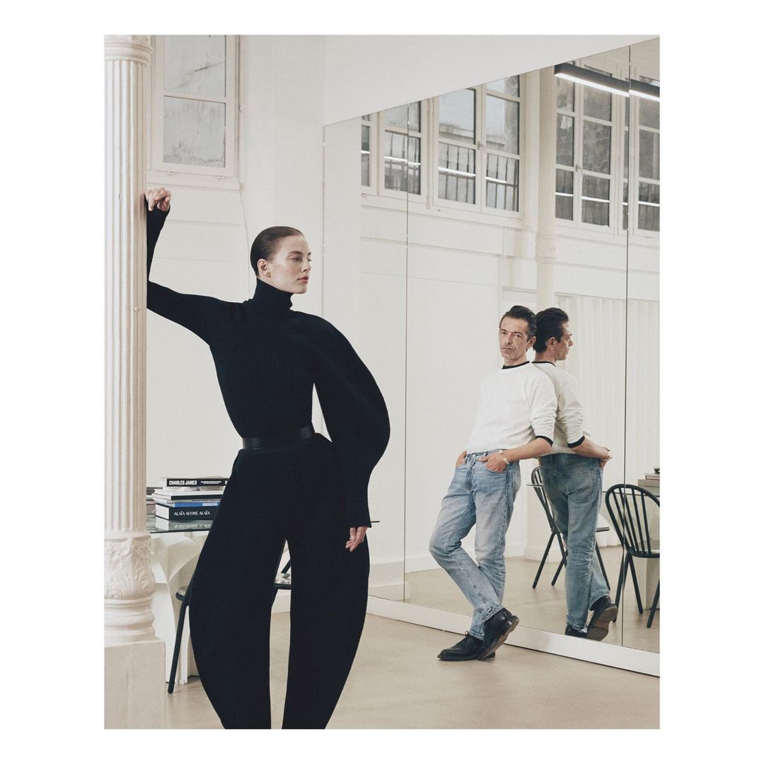 Fashion shot of a slender woman in a black outfit posing in a white studio space alongside a man in jeans and a white top.