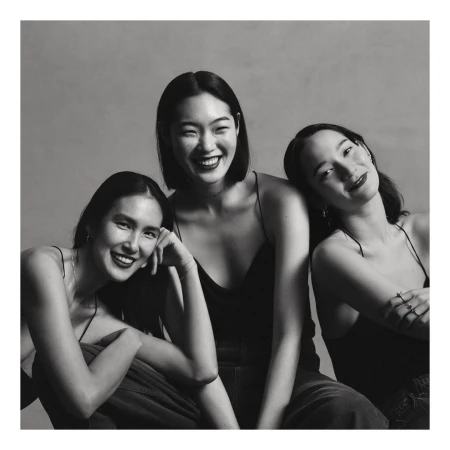 Three smiling women in dark tops posing together against a neutral studio background in a black and white portrait.