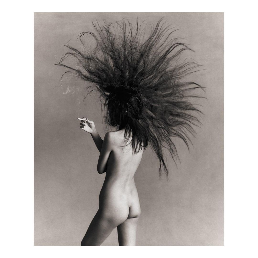 Back view of a topless woman with long black hair smoking a cigarette in a photo studio with a solid grey background in black and white.