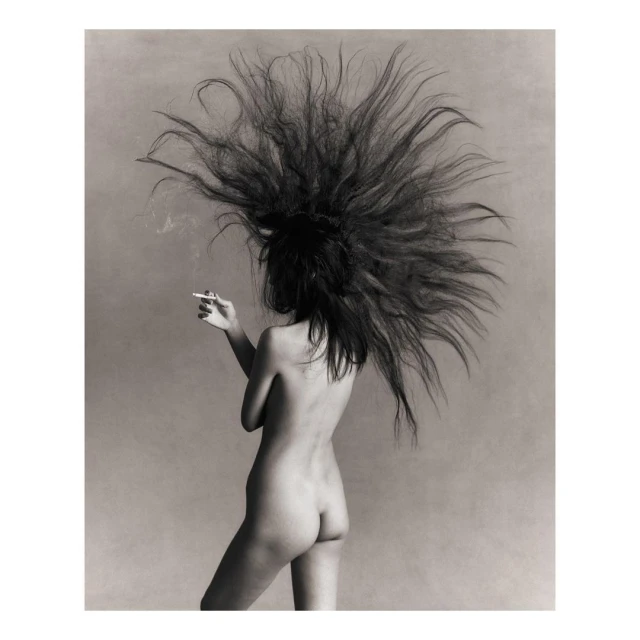 Back view of a topless woman with long black hair smoking a cigarette in a photo studio with a solid grey background in black and white.