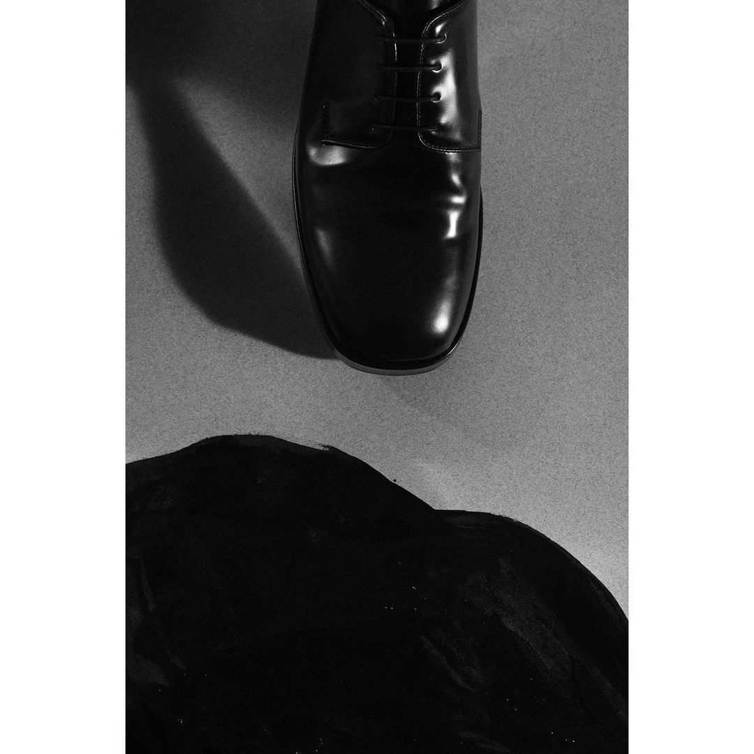Black leather dress shoe with glossy finish on gray backdrop with diffused lighting and dark fluid shape, creating dramatic monotone.