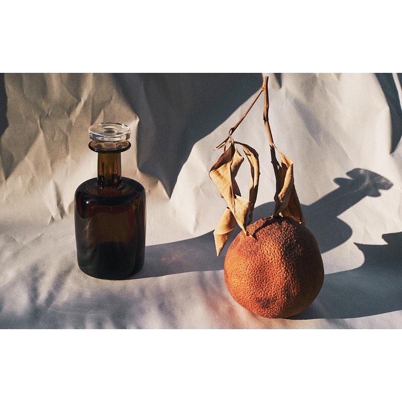 Still life featuring a brown glass bottle and a dried orange with a branch, set against a softly wrinkled white background.
